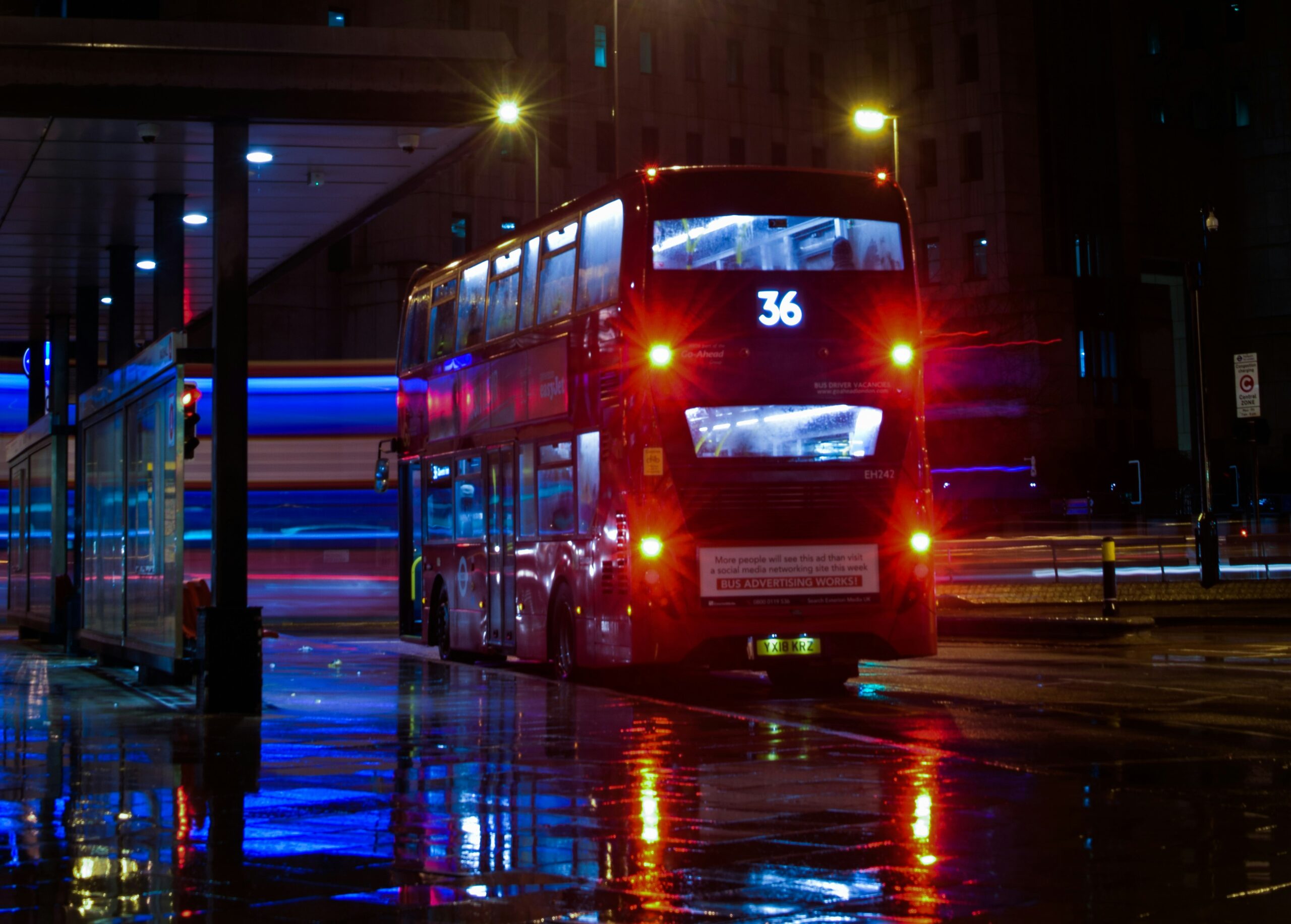 Nighttime view of Humberston Bus Station with dim streetlights casting a soft glow over empty bus bays. A few buses are parked, their headlights off, while the station is quiet and deserted. The dark sky contrasts with the illuminated signs and reflective surfaces on the buses.
