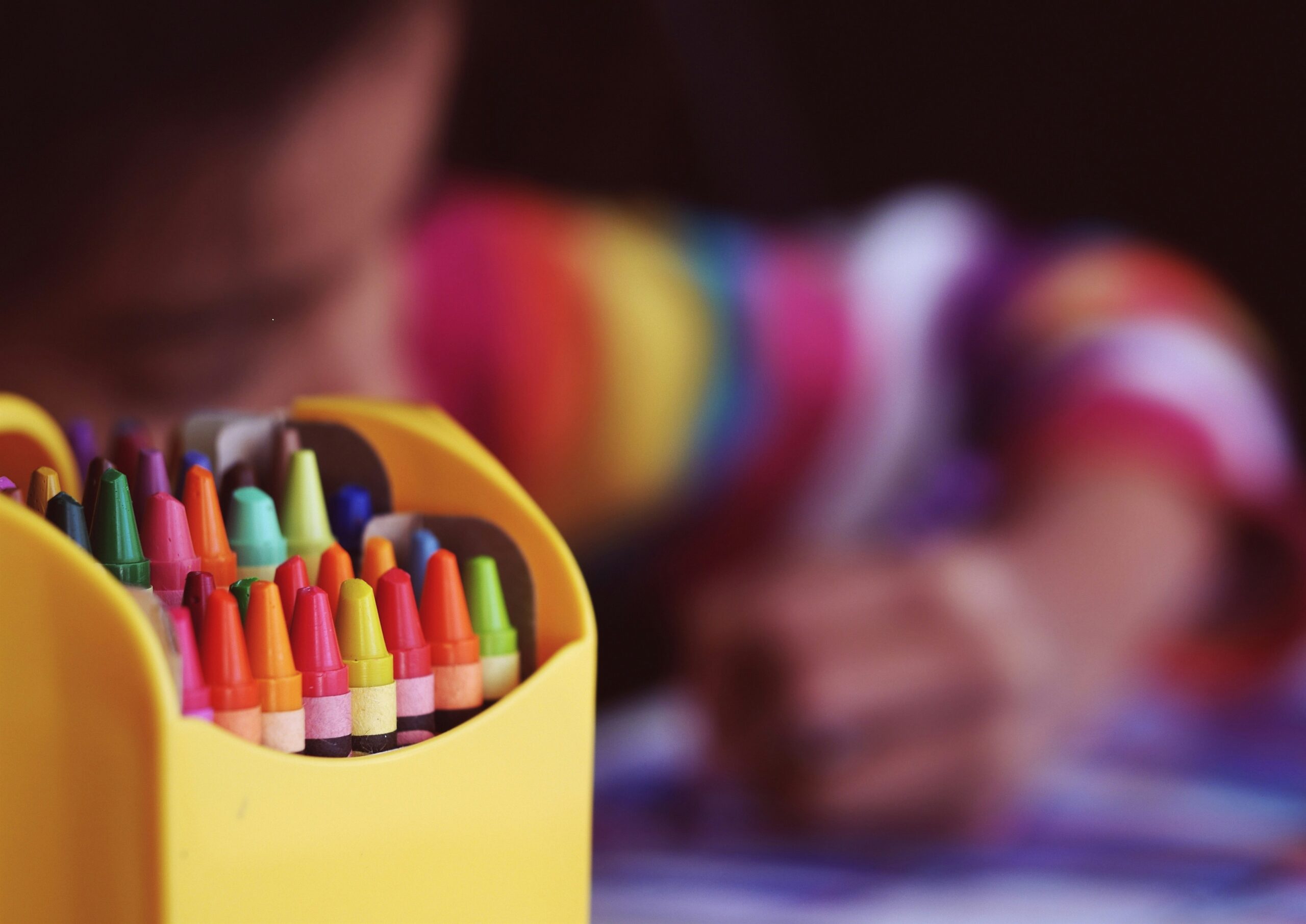 Close-up of a box of colorful crayons with a blurred child in the background, focusing on drawing while wearing a rainbow-striped sweater.